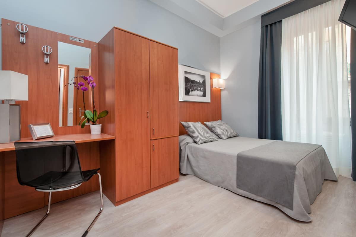 A hotel room with a wooden wardrobe, a desk with a chair, a bed with a grey bedspread, a framed picture on the wall, and a window with curtains.