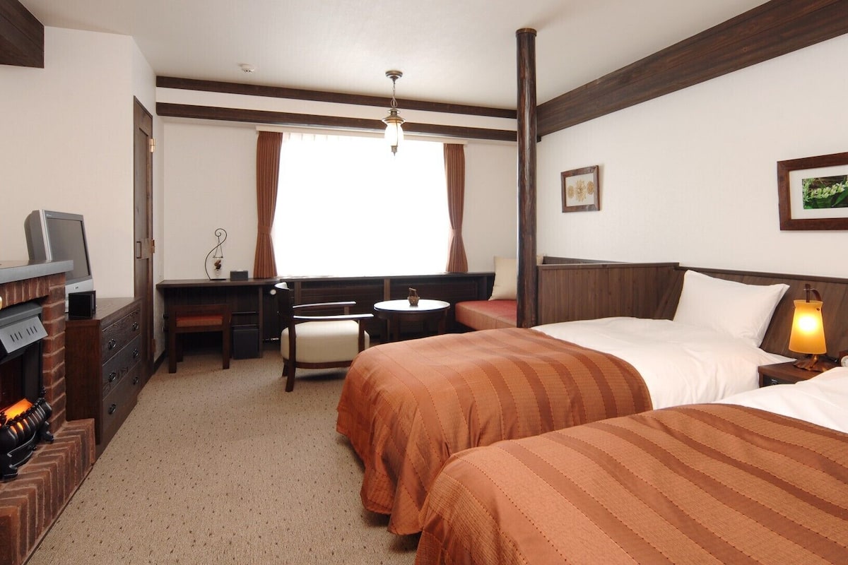 A hotel room with two beds, a desk, a chair, a fireplace, and a window with curtains.