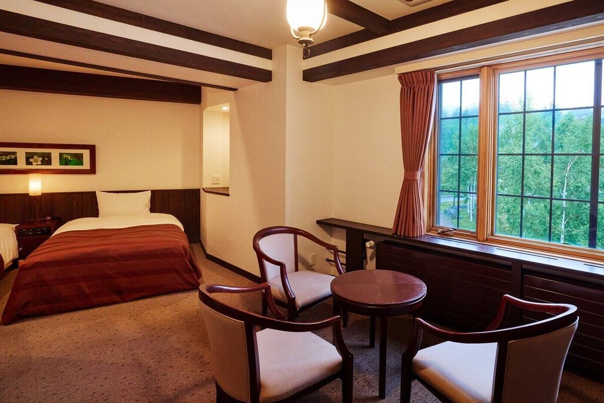 Une chambre d’hôtel avec un lit, deux chaises, une petite table et une fenêtre donnant sur un paysage verdoyant.