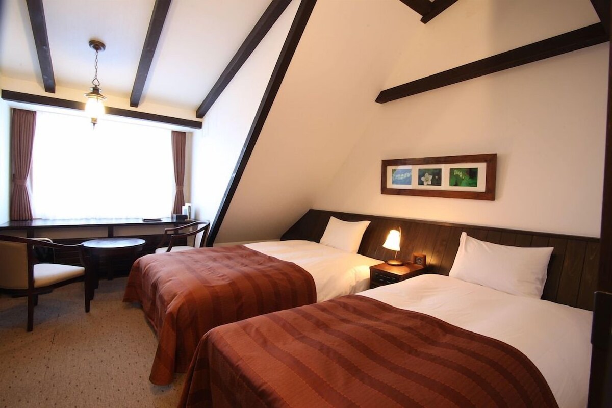 Habitación de hotel con dos camas, un escritorio, una silla, una lámpara y un cuadro en la pared.