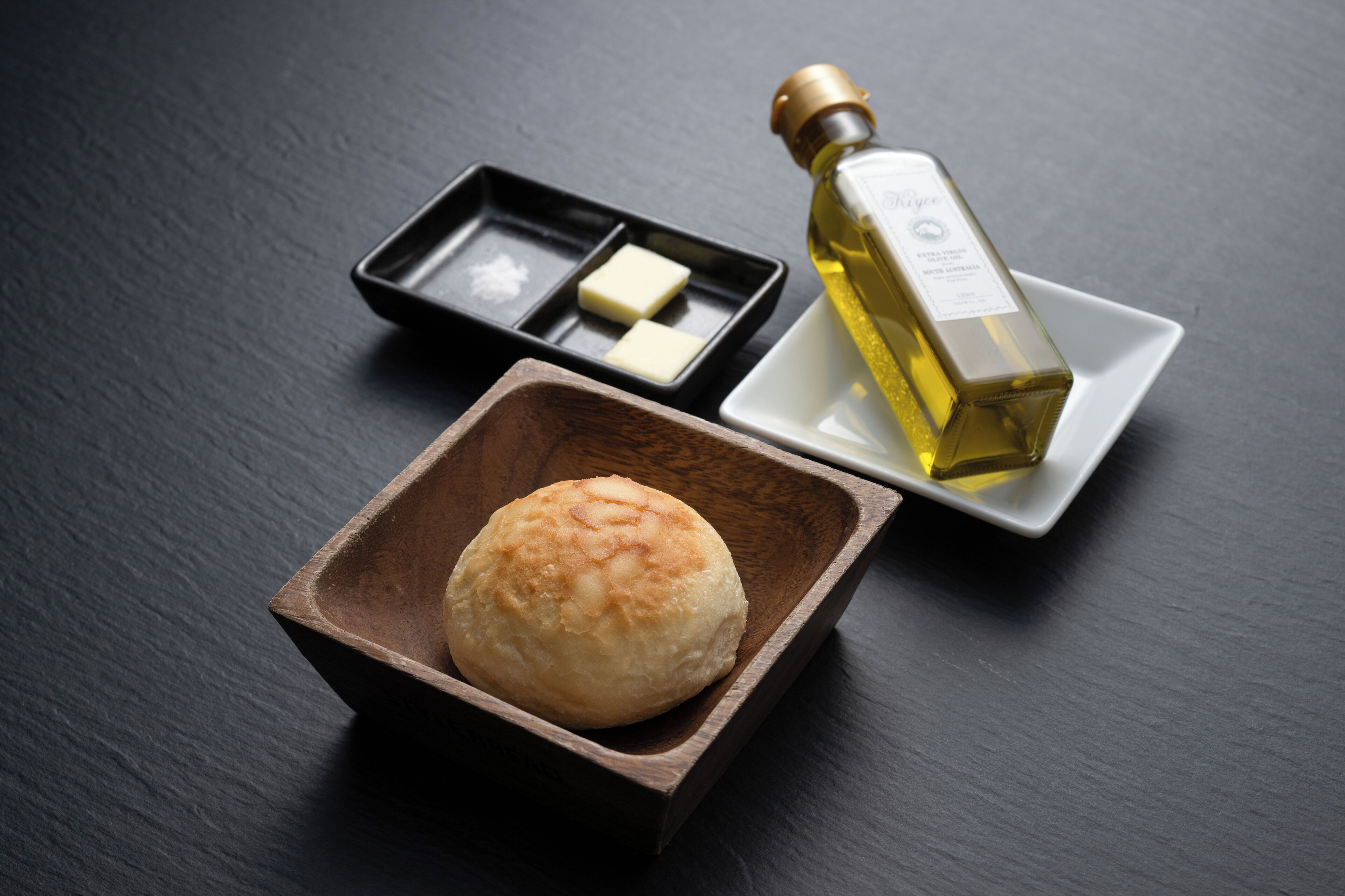 A bottle of olive oil, a small wooden bowl with a bun, and a black tray with two pieces of butter on a dark surface.