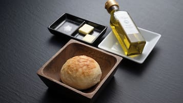 A bottle of olive oil, a small wooden bowl with a bun, and a black tray with two pieces of butter on a dark surface.