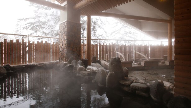 Un onsen japonés tradicional con bañeras termales de piedra, barandillas de madera y un fondo de pared de piedra.