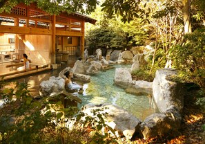 A traditional Japanese onsen with a wooden building, a clear hot spring surrounded by rocks, and a natural forested area.