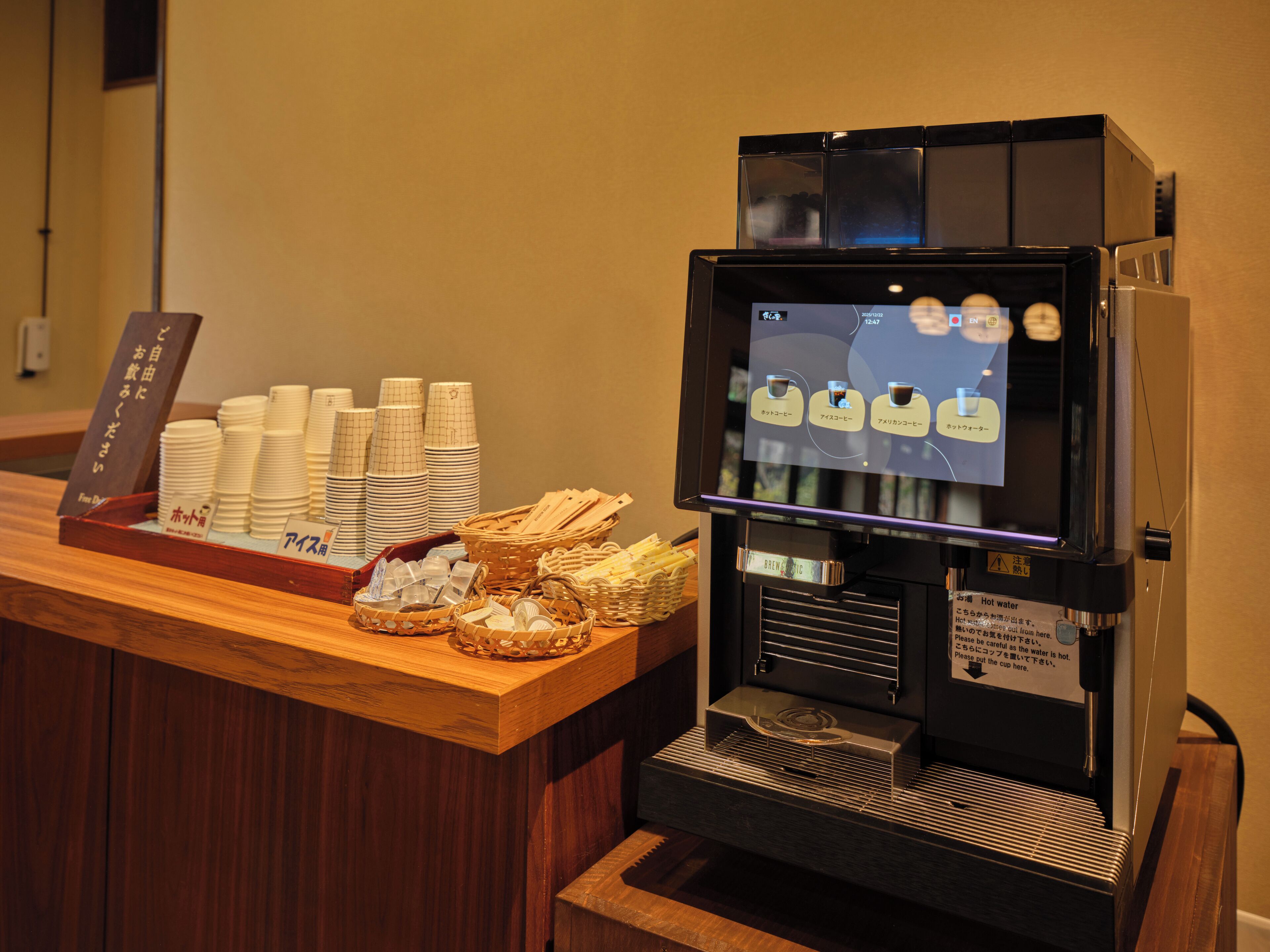 A coffee machine with a digital menu, stacked paper cups, and a wooden counter.