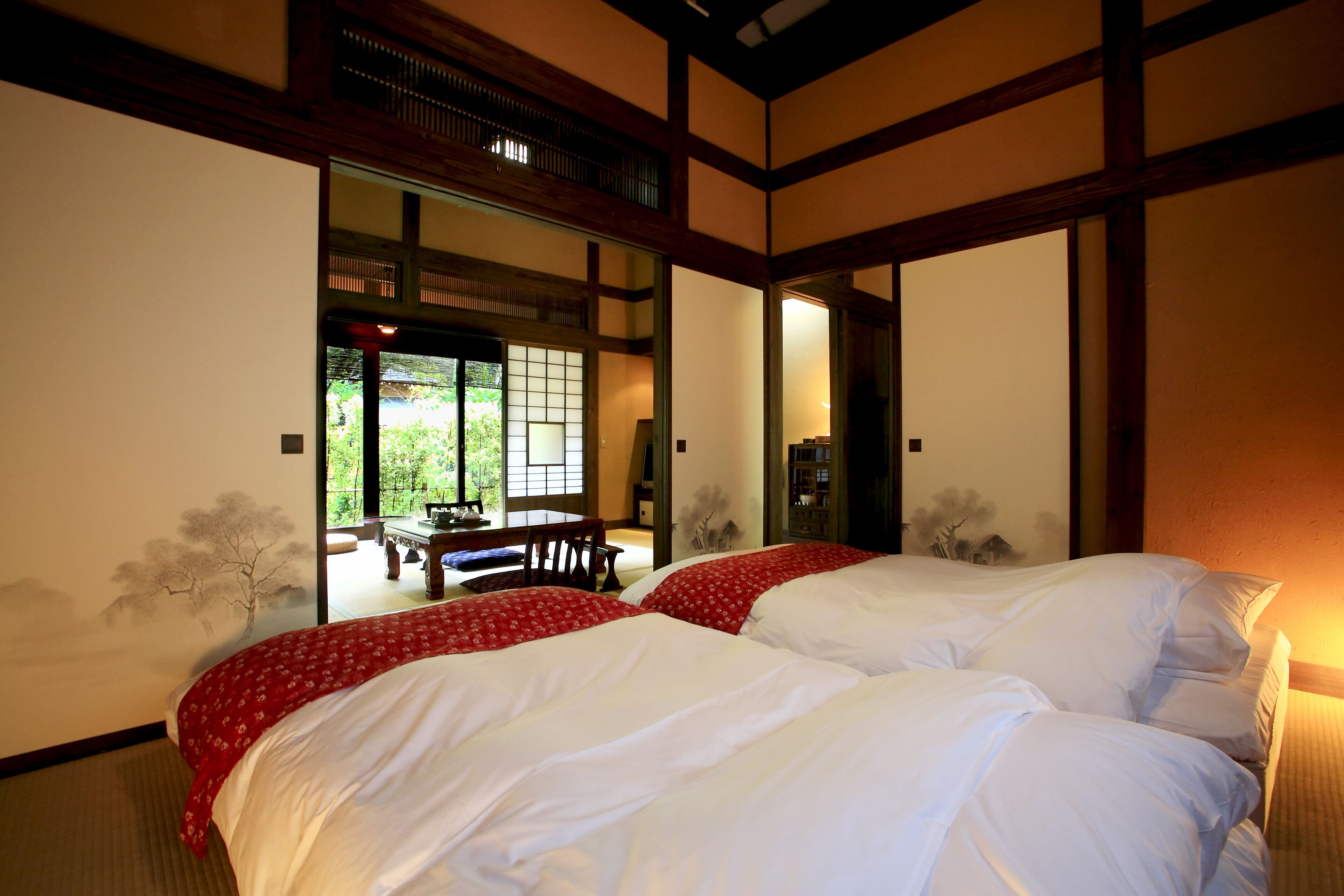 A traditional Japanese-style bedroom with two beds, a sliding door leading to a dining area, and a view of greenery outside.