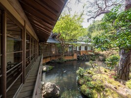 A traditional Japanese building with a wooden deck overlooking a serene pond, surrounded by lush greenery and stone lanterns.