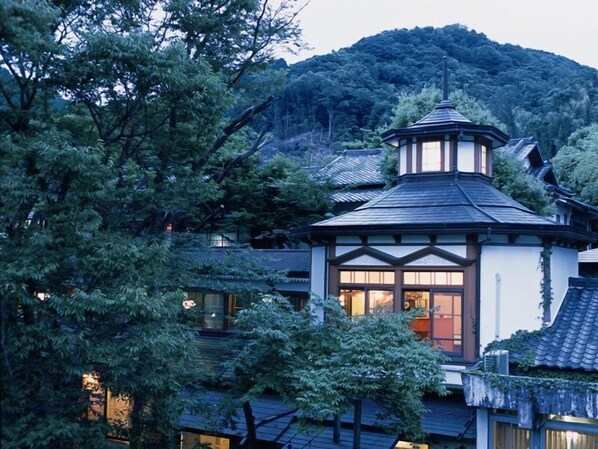 Un edificio de estilo japonés tradicional con una imponente torre central, rodeado de exuberante vegetación y un telón de fondo montañoso.