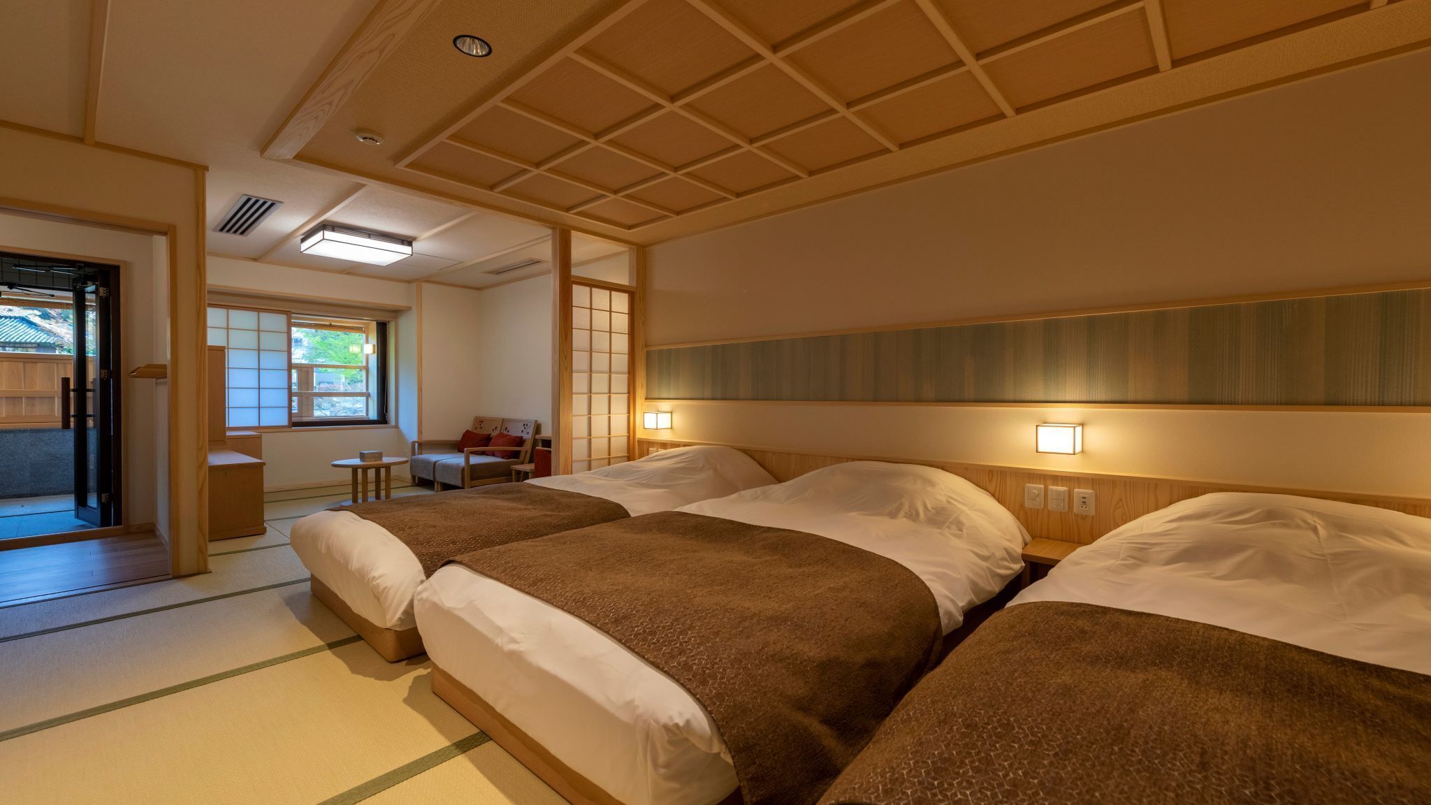 A hotel room with two beds, a sitting area with a chair and table, and a view of the outside.