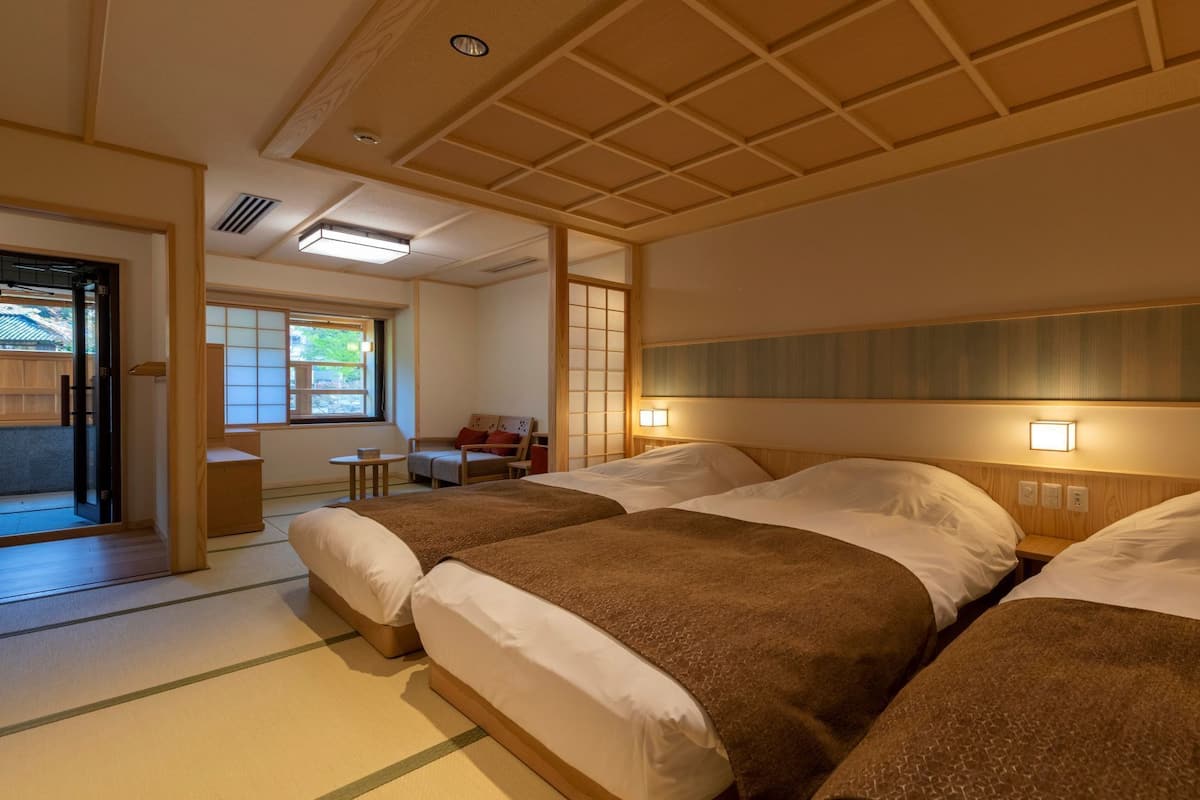 Habitación de hotel con dos camas, una zona de estar con una silla y una mesa, y vistas al exterior.