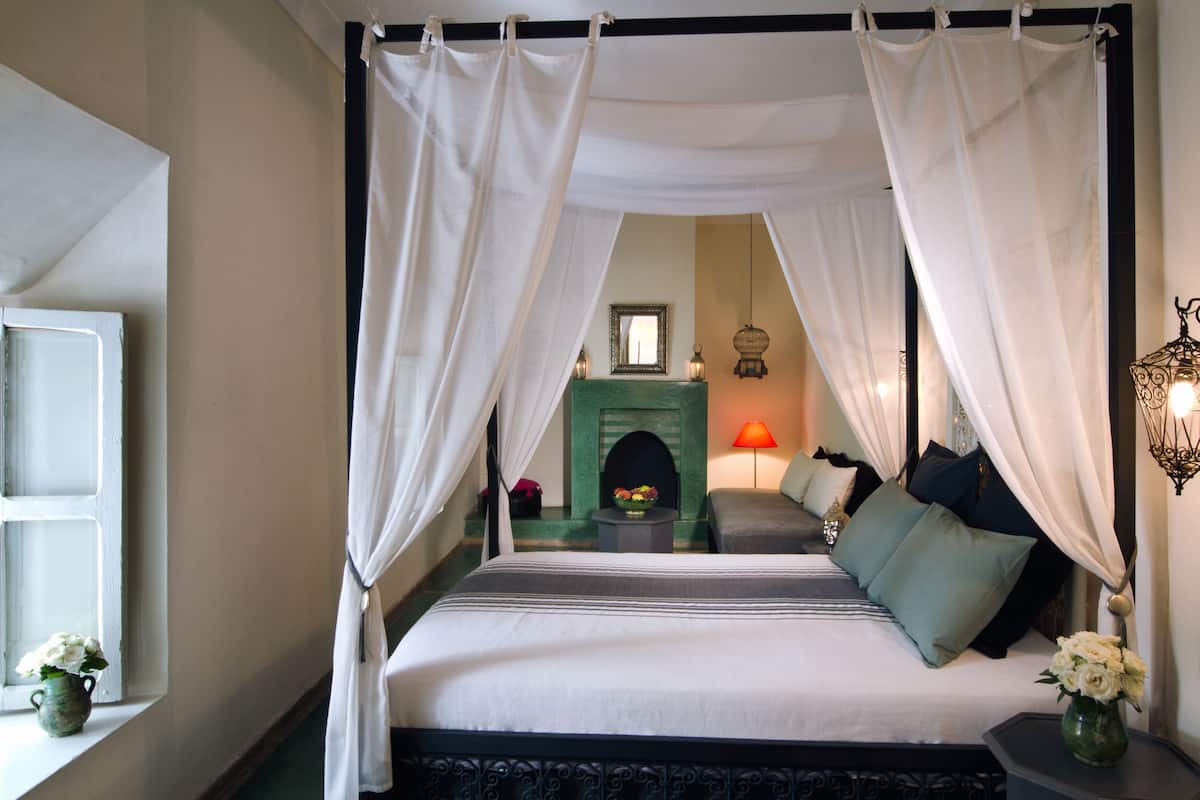 Uma cama de dossel com um manto verde, uma mesa de cabeceira com abajur e um vaso de flores.