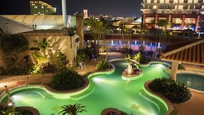 A swimming pool with a winding design, surrounded by plants and benches, set against a backdrop of buildings and a night sky.