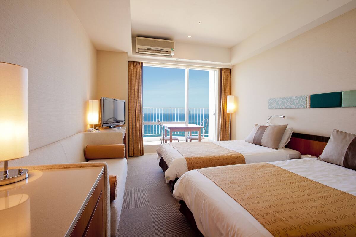 A hotel room with two beds, a desk, a chair, and a balcony with a view.