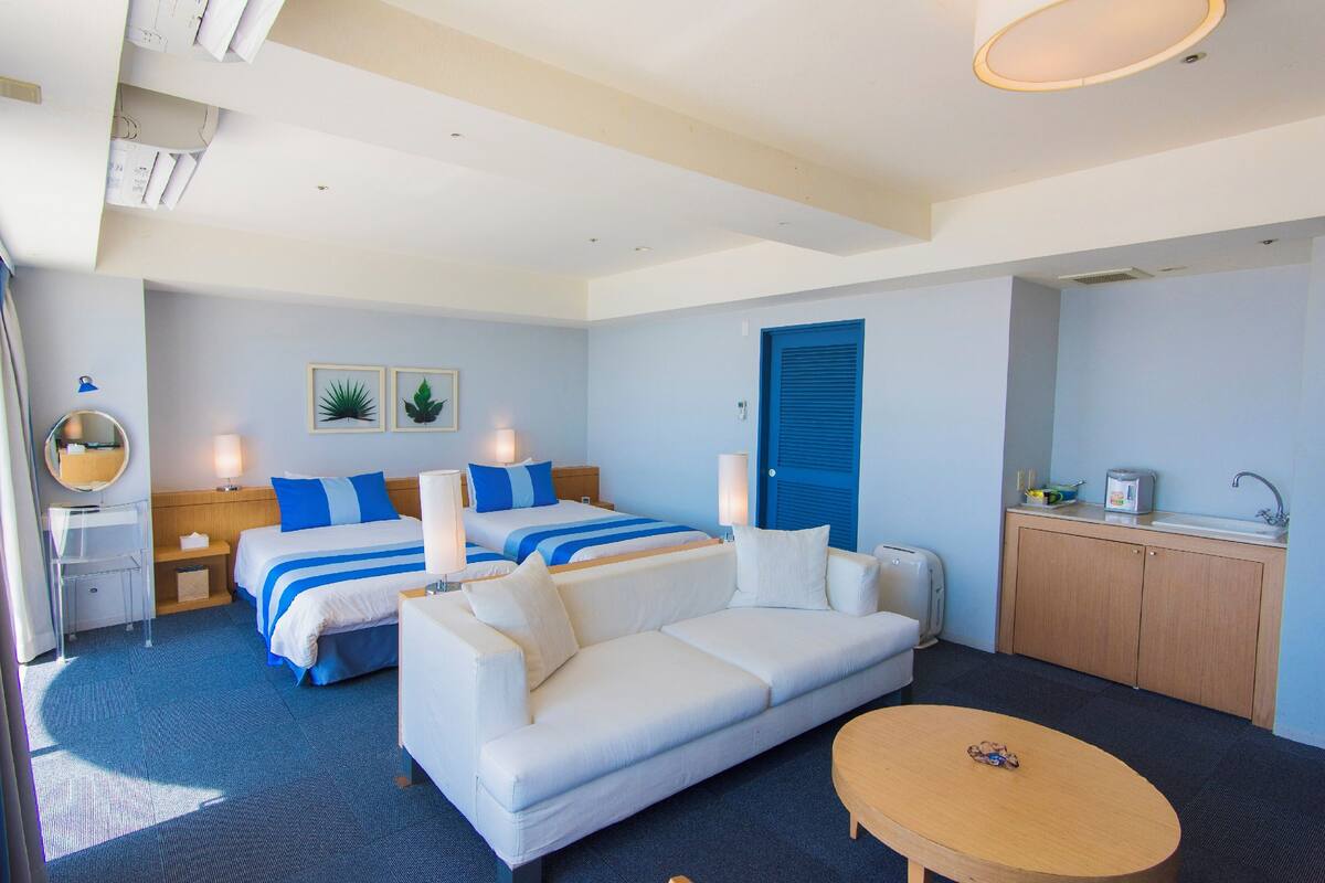 A hotel room with two beds, a sofa, a small table, a desk with a sink, and a mirror.