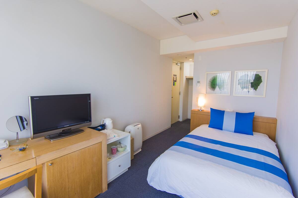A hotel room with a bed, a desk with a television, a small table with a lamp, and a door leading to another room.