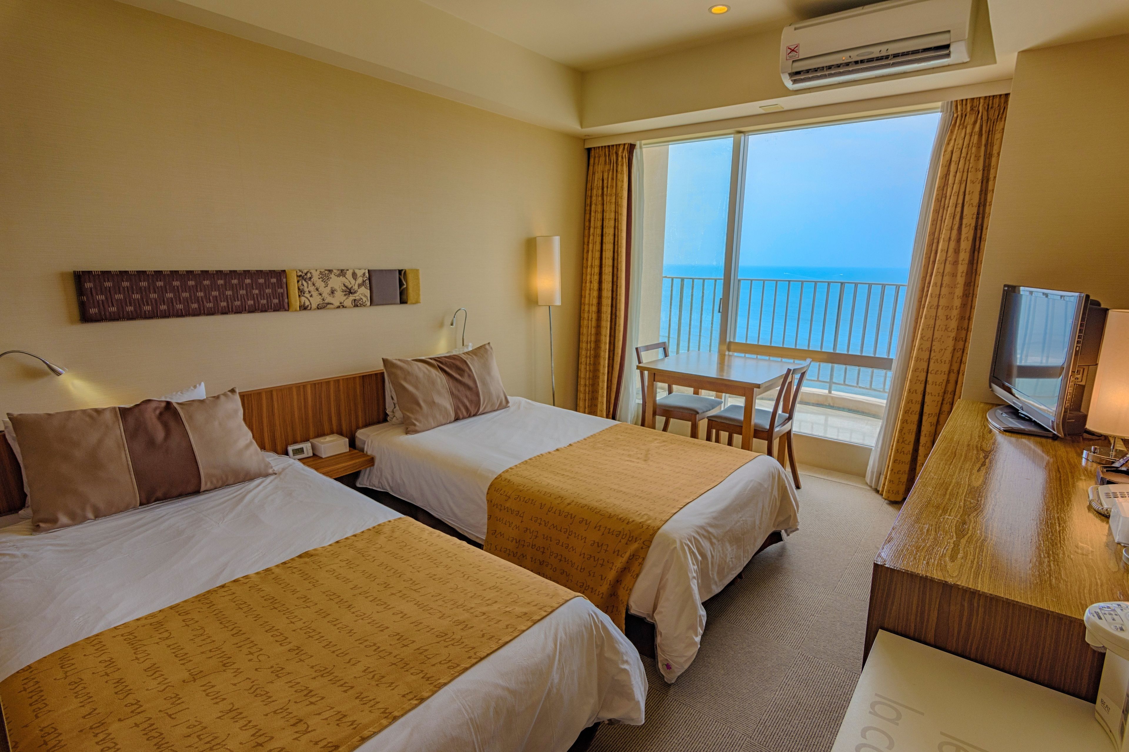 A hotel room with two beds, a desk, a chair, a TV, and a balcony with a view of the sea.
