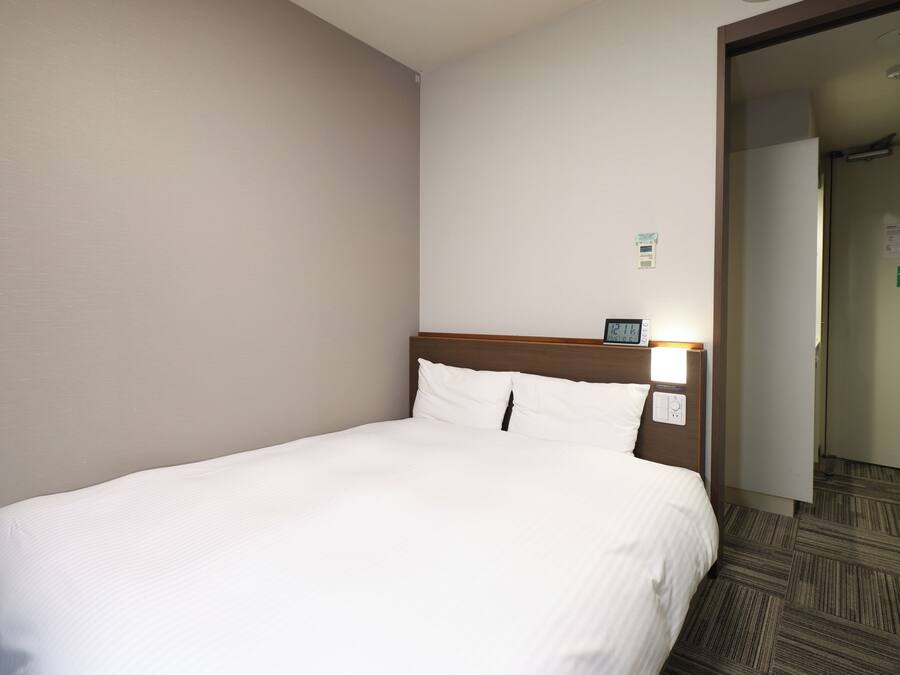 A hotel room with a single bed, a wooden headboard, a nightstand with a clock, and a carpeted floor.
