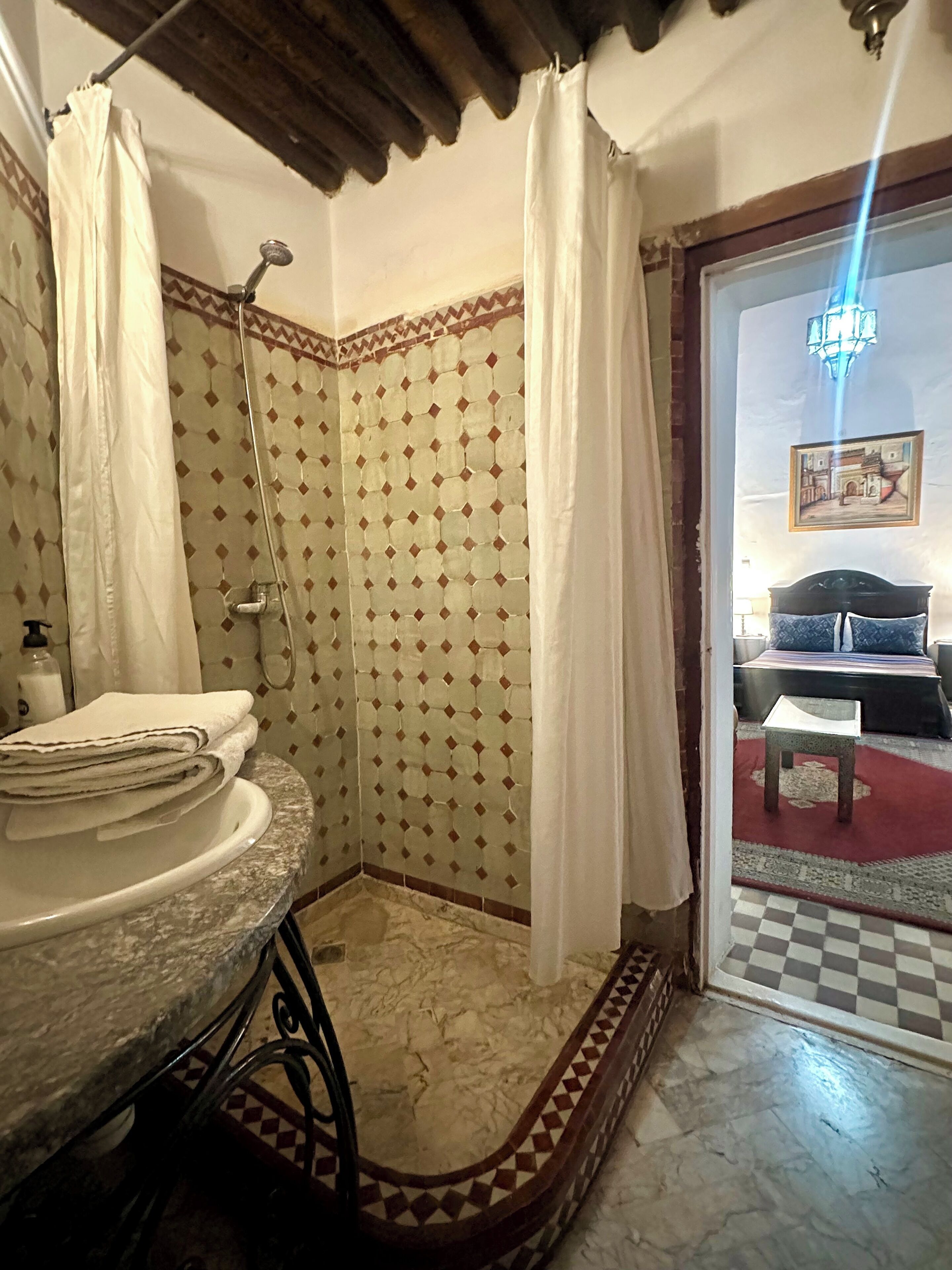 Une salle de bain avec un lavabo, une douche et un lit visible à travers une porte ouverte.