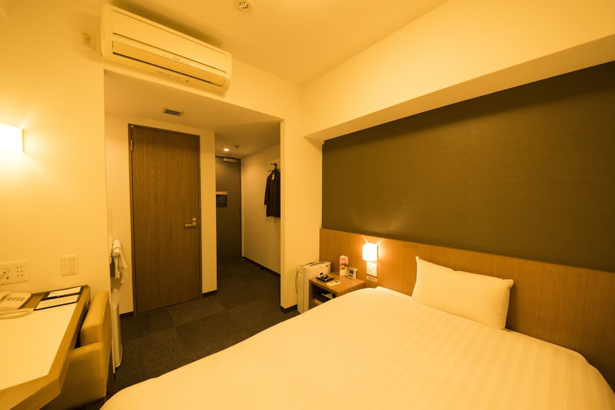 A hotel room with a bed, a desk, a chair, a nightstand, a lamp, and a door.