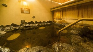 Un onsen japonès tradicional amb una banyera de pedra envoltat de roques, una banca de fusta i una paret de fusta amb una placa.
