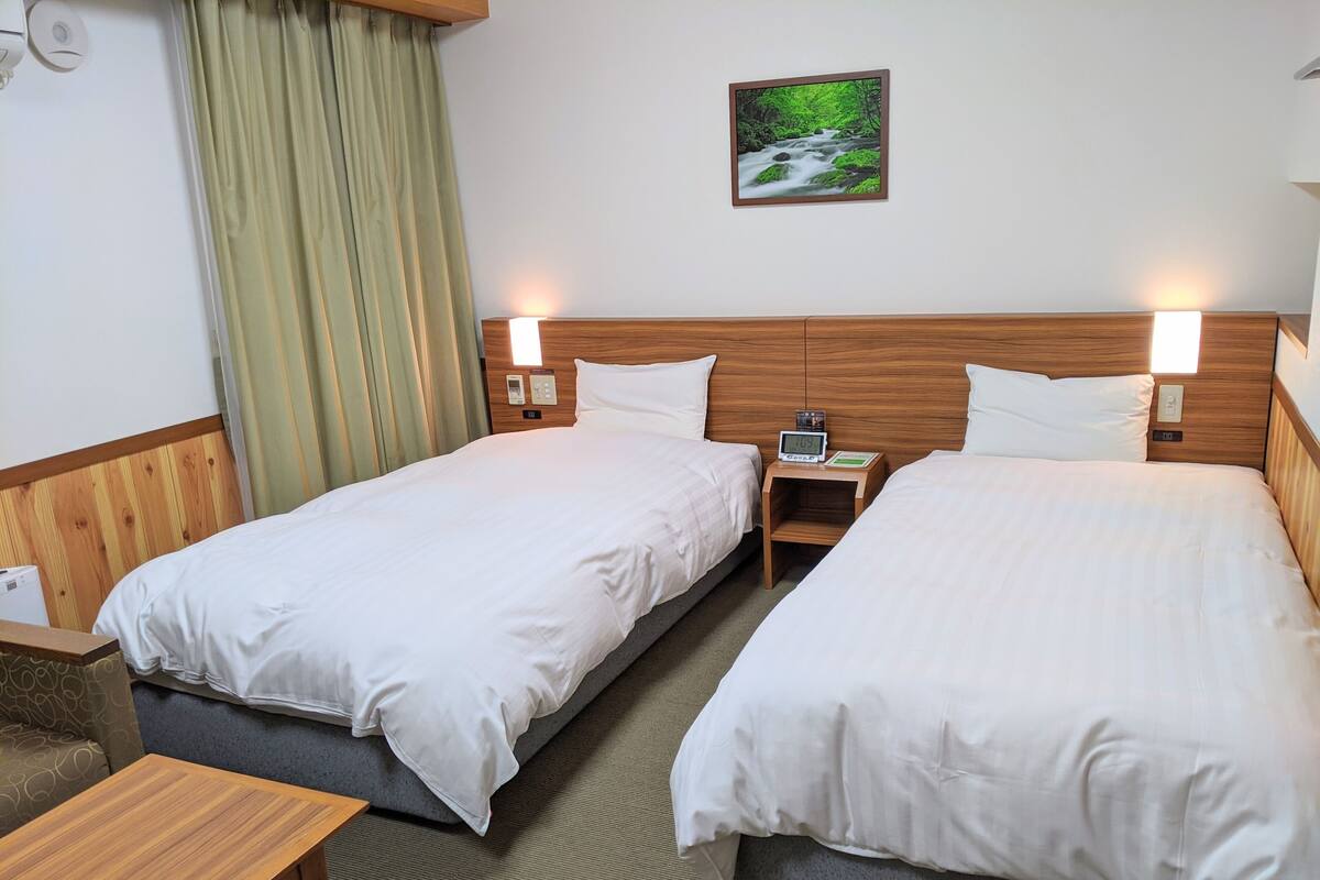 A hotel room with two beds, wooden headboards, a small bedside table with a clock, a wall-mounted painting, and a window with curtains.