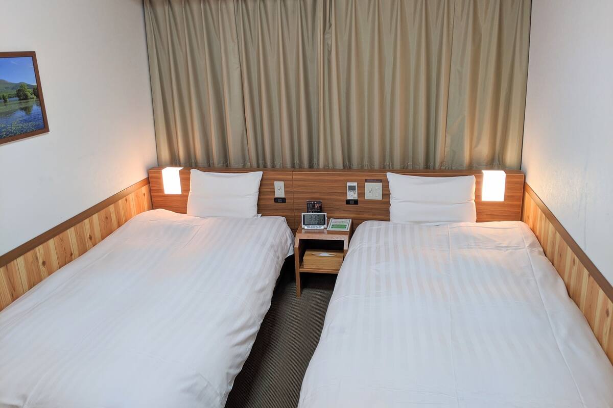 A hotel room with two beds, a nightstand, a wall-mounted clock, and a window with curtains.