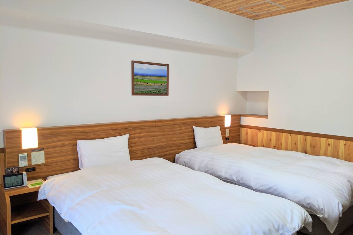 A hotel room with two single beds, wooden headboards, bedside lamps, a nightstand with a clock, and a framed picture on the wall.