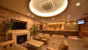 A lobby with a stone fireplace, comfortable seating, a flat-screen TV, and a digital screen displaying information. - Dormy Inn PREMIUM Sapporo Hot Spring (Sapporo)