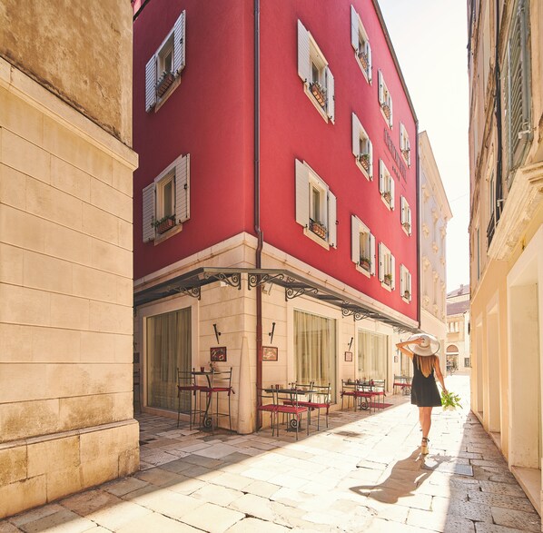 Une rue étroite bordée d’un immeuble rouge, doté de nombreuses fenêtres avec volets et d’une terrasse.
