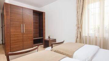 Quarto de hotel com guarda-roupa de madeira, duas camas com roupa de cama bege e branca, uma pequena mesa de cabeceira e uma janela com cortinas finas.