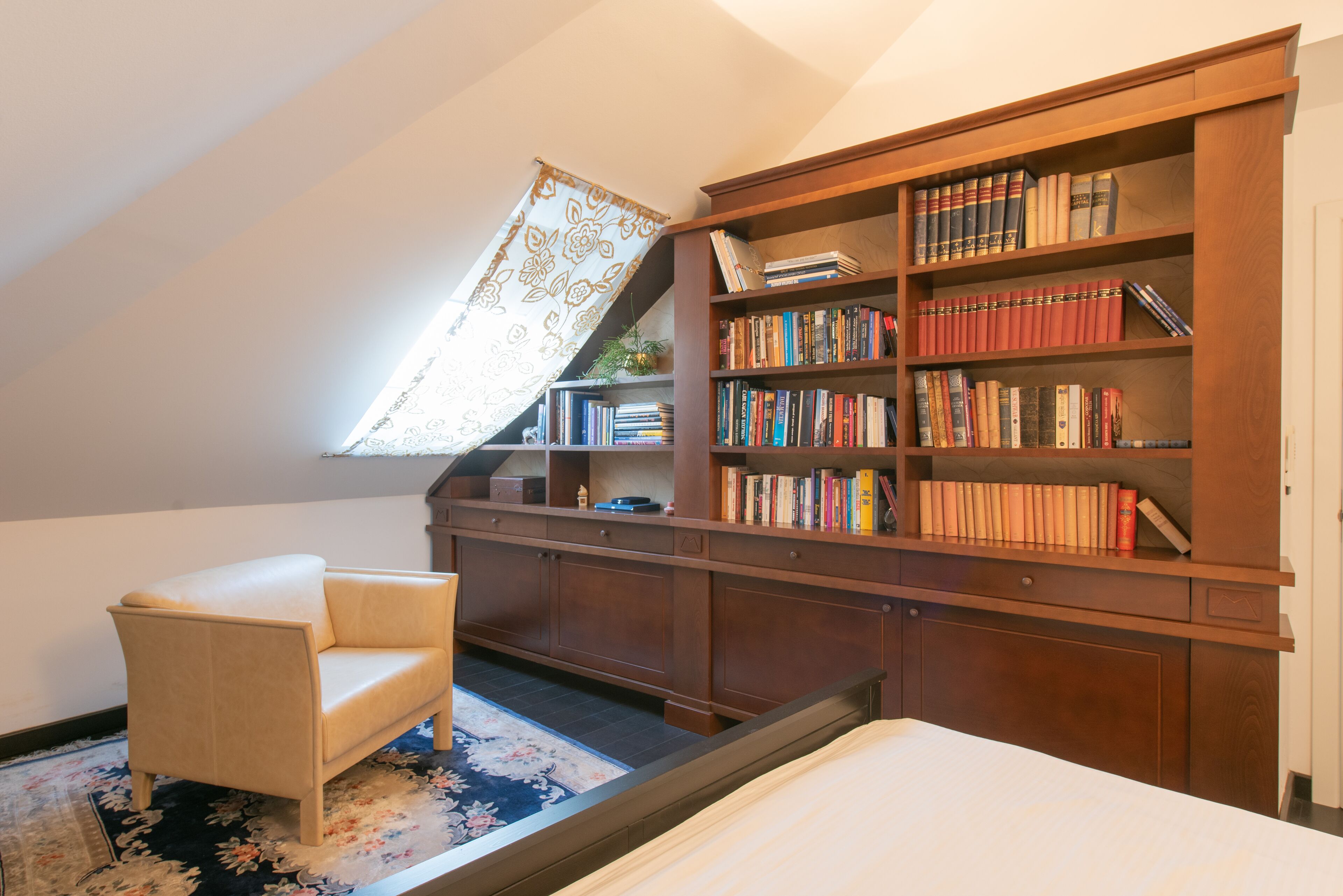 A cozy attic bedroom with a built-in wooden bookshelf, a comfortable chair, and a slanted ceiling allowing natural light.