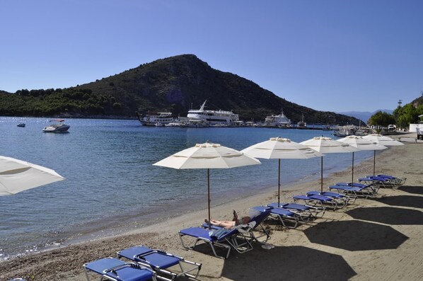 A beach with rows of sun umbrellas and lounge chairs, boats docked in the background, and a mountainous landscape.