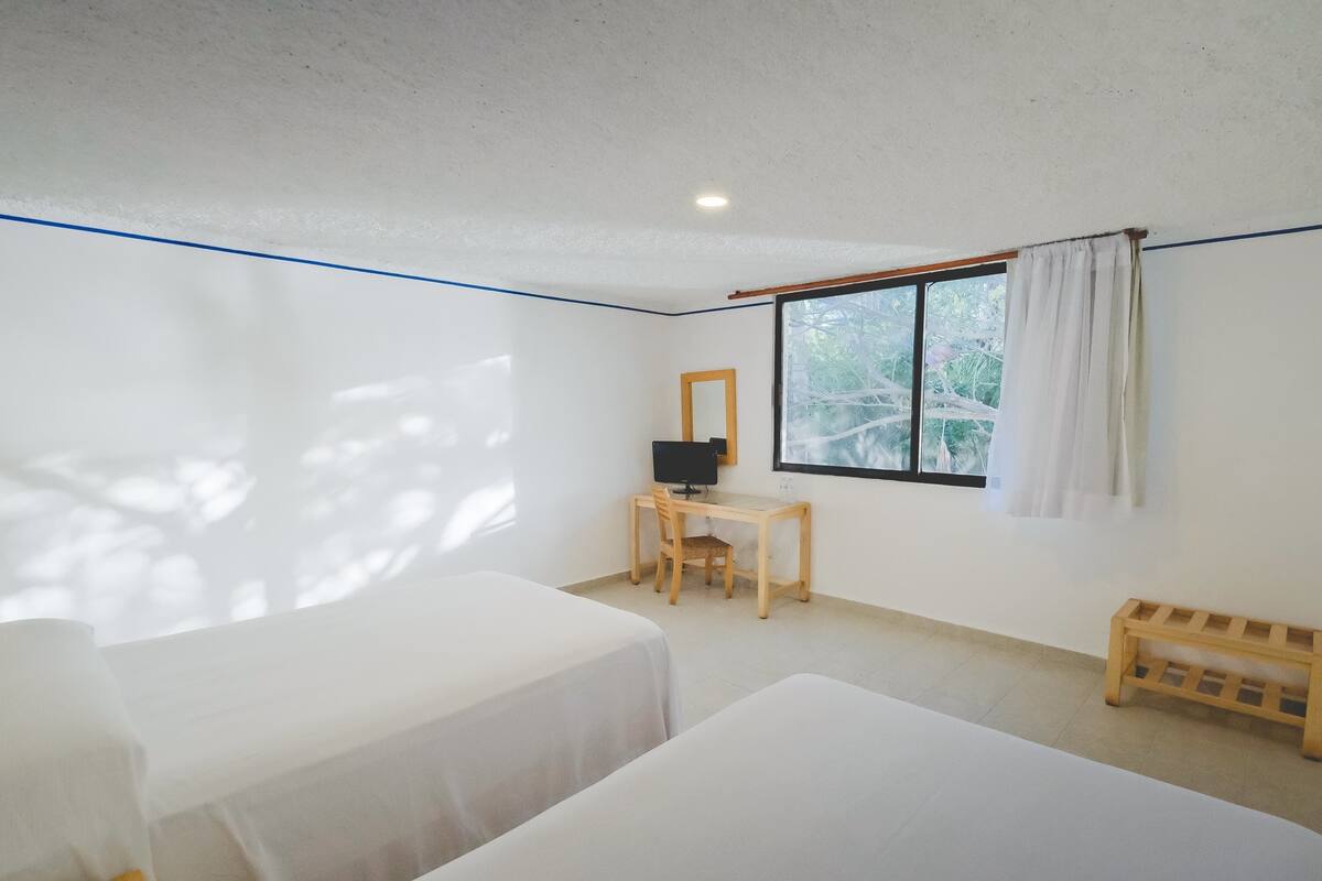 Una habitación con dos camas, un escritorio con computadora, una ventana que da a vistas de árboles y un banco de madera.