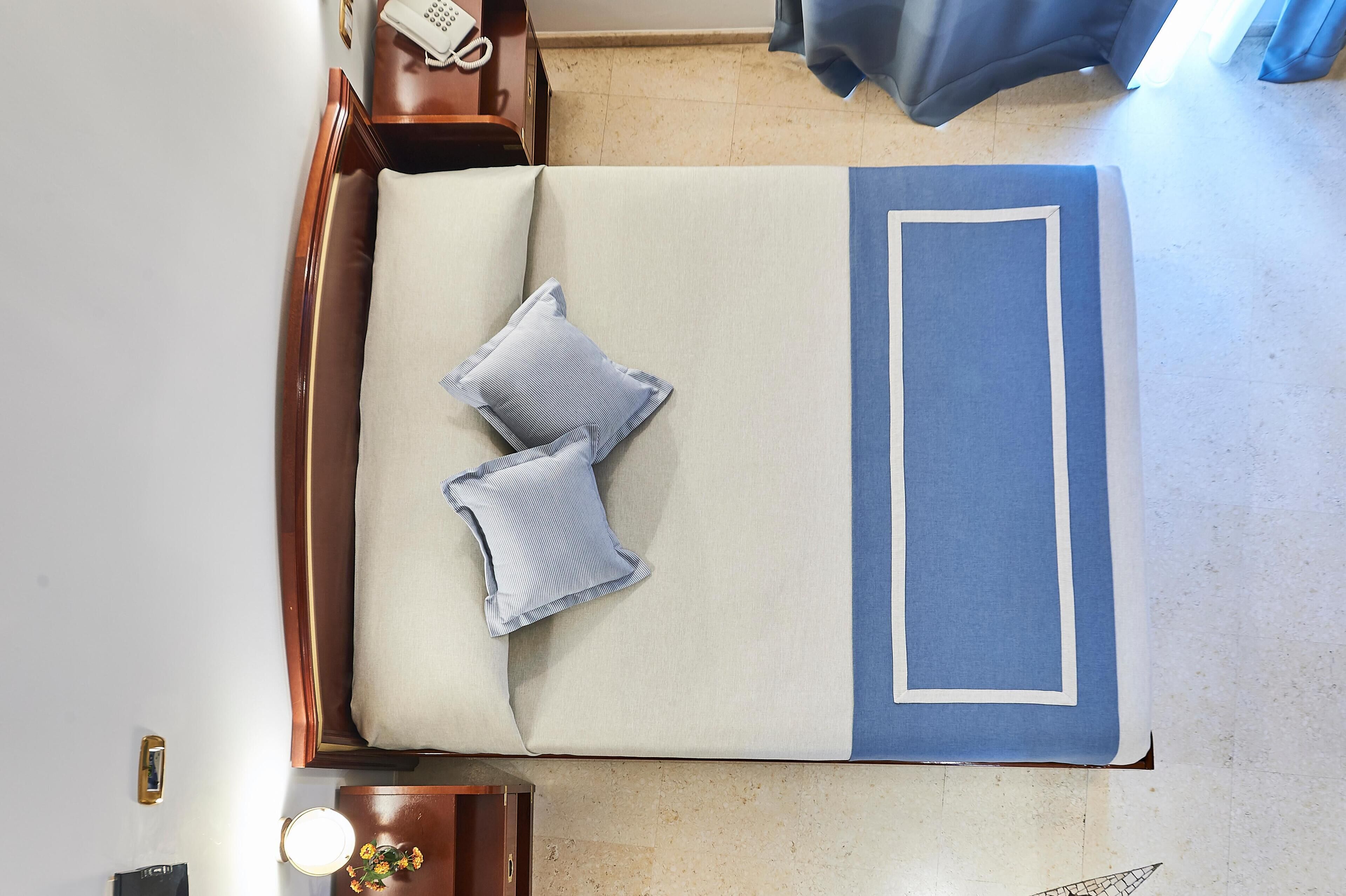 Una cama con una colcha azul y blanca, dos almohadas, una mesita de noche con un teléfono y una pequeña mesa con una lámpara.