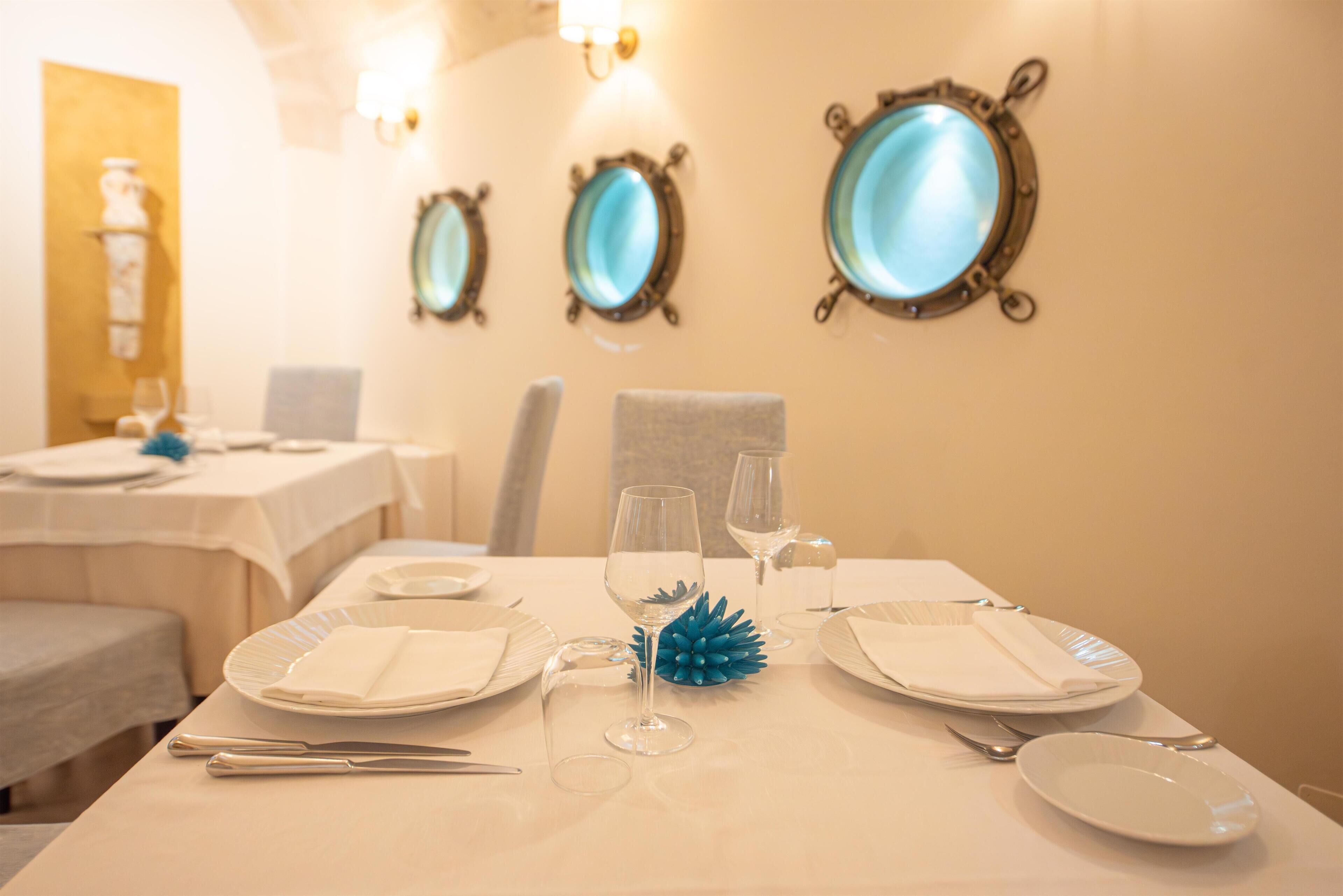 Una mesa de comedor dispuesta con platos, cubiertos y un objeto decorativo azul, con espejos montados en la pared y un reloj al fondo.