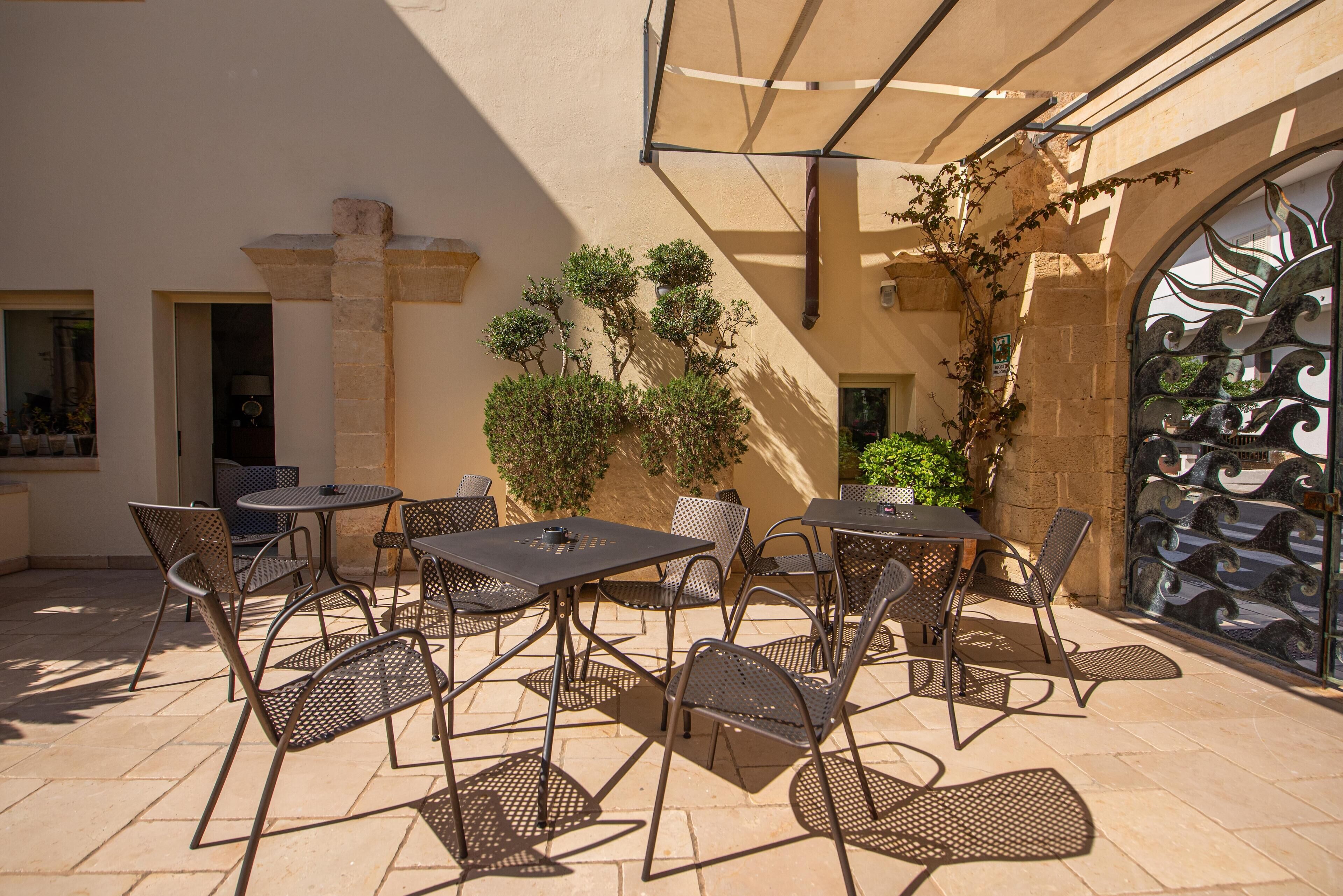 Un patio al aire libre con mesas y sillas de metal, rodeado de plantas y una puerta de metal decorativa.