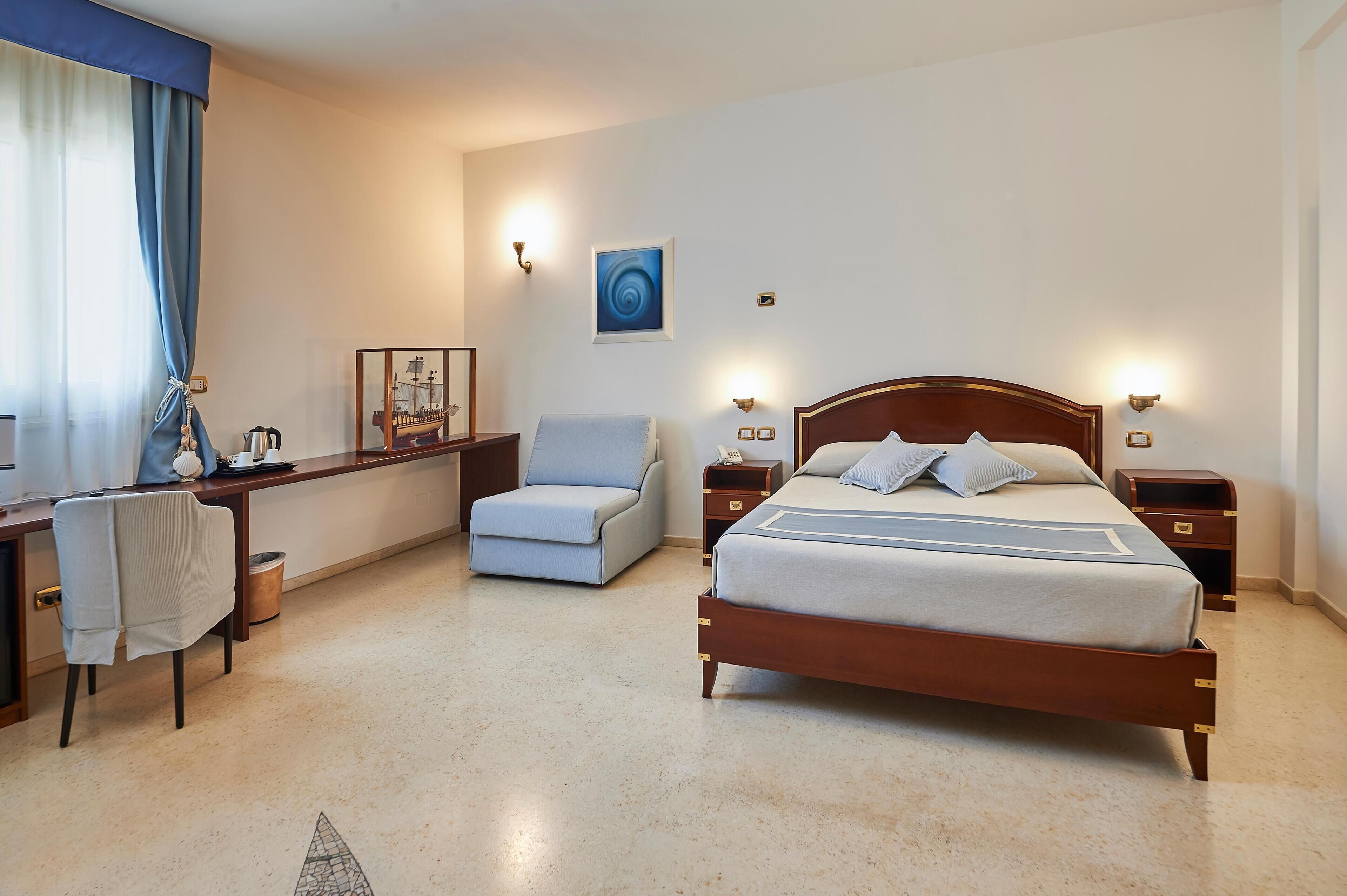 A hotel room with a bed, a desk, a chair, and a small table with a lamp.