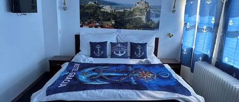 Quarto de hotel com cama, colcha temática de âncora, cortina com tema náutico e televisão fixada na parede.