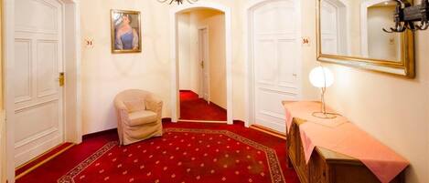 Un couloir avec un tapis rouge, un fauteuil beige, un miroir et une table avec une lampe.