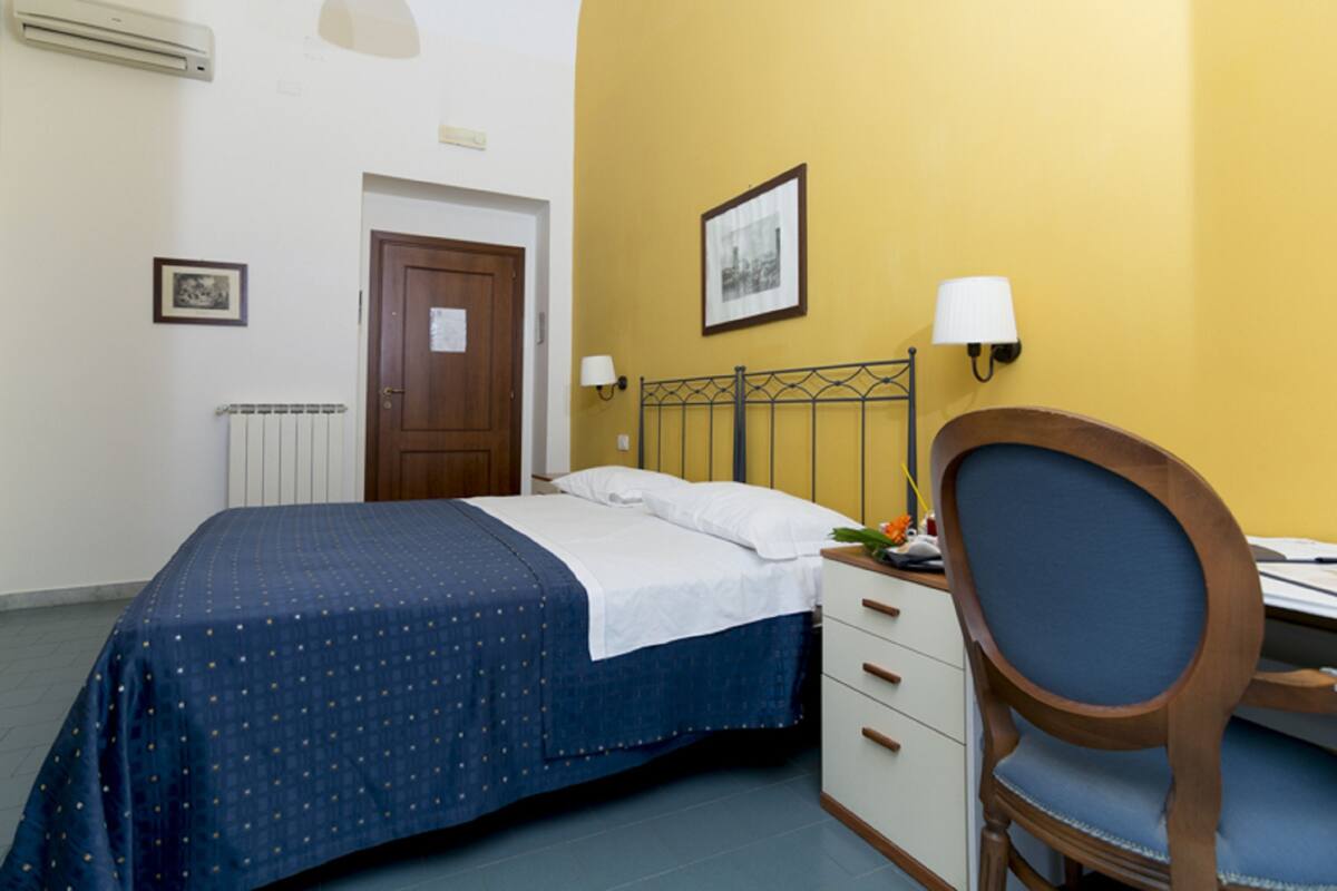 A bedroom with a bed, a desk, a chair, a radiator, a door, and a wall-mounted lamp.