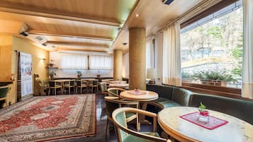 A hotel lobby with a large central rug, multiple seating areas including a round table and a sofa, and a view of a garden through large windows.