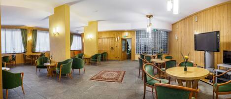 Een hotel lobby met groene stoelen, houten tafels, een televisie en een groot raam met gordijnen.