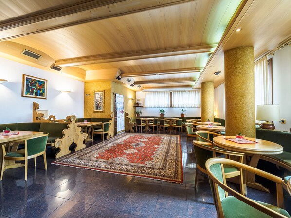 A spacious restaurant with wooden ceiling beams, green upholstered seating, and a large patterned rug on the floor. - Hotel Sant Anton (Bormio)