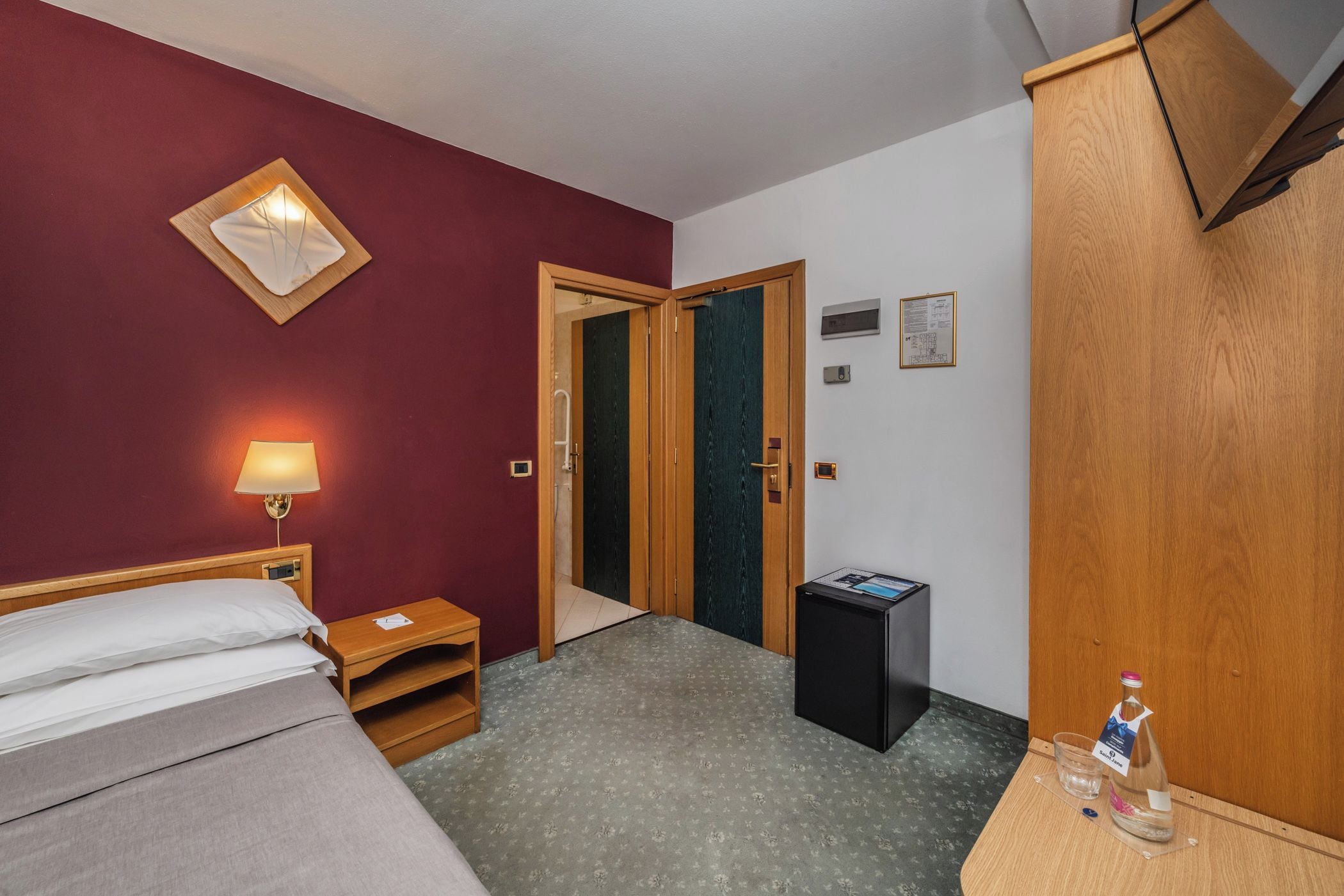 A hotel room with a bed, bedside table, lamp, wardrobe, and a small trash bin.
