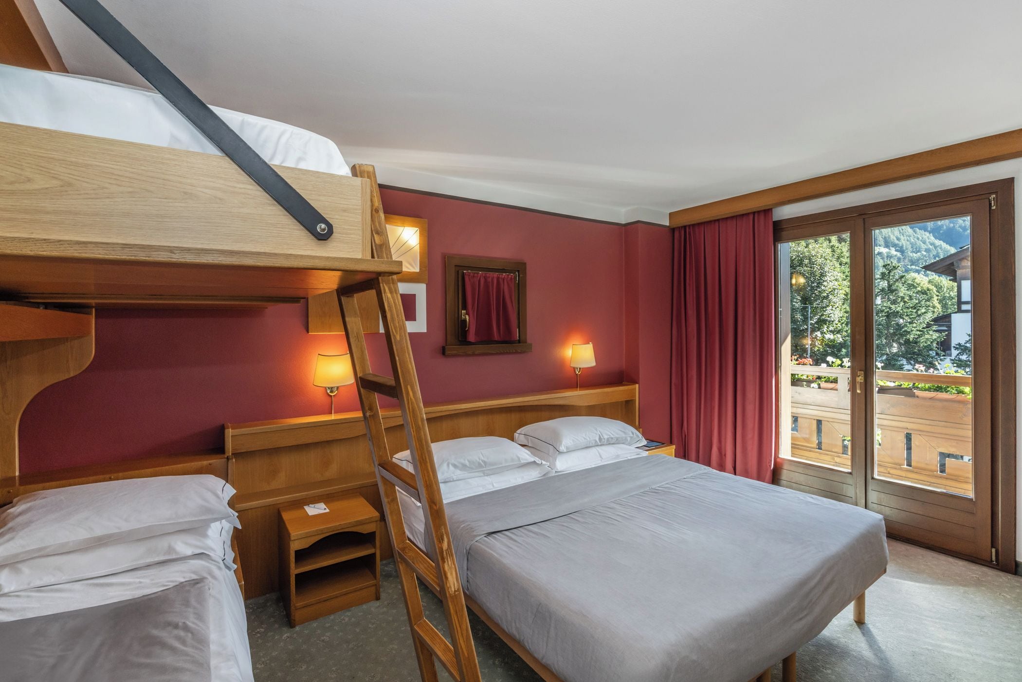 Une chambre d’hôtel avec un lit superposé, un lit simple, une échelle en bois, une table de chevet, une lampe et une vue sur un balcon donnant sur un paysage extérieur.