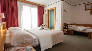 A hotel room with two beds, a wooden desk, a chair, a radiator, and a window with curtains.