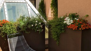 A modern outdoor seating area with a metal chair and a glass-top table, surrounded by flower planters and a wooden deck. - Citta Trüffel Hotel (Wiesbaden)