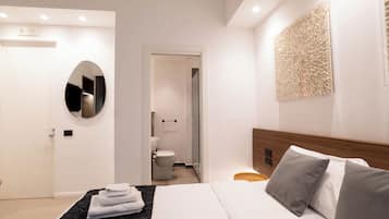 Ein modernes Hotelzimmer mit einem ordentlich bezogenen Bett, einem runden Spiegel, einem Holzkopfende und einem durchgegangene Tür erkennbaren Badezimmer.