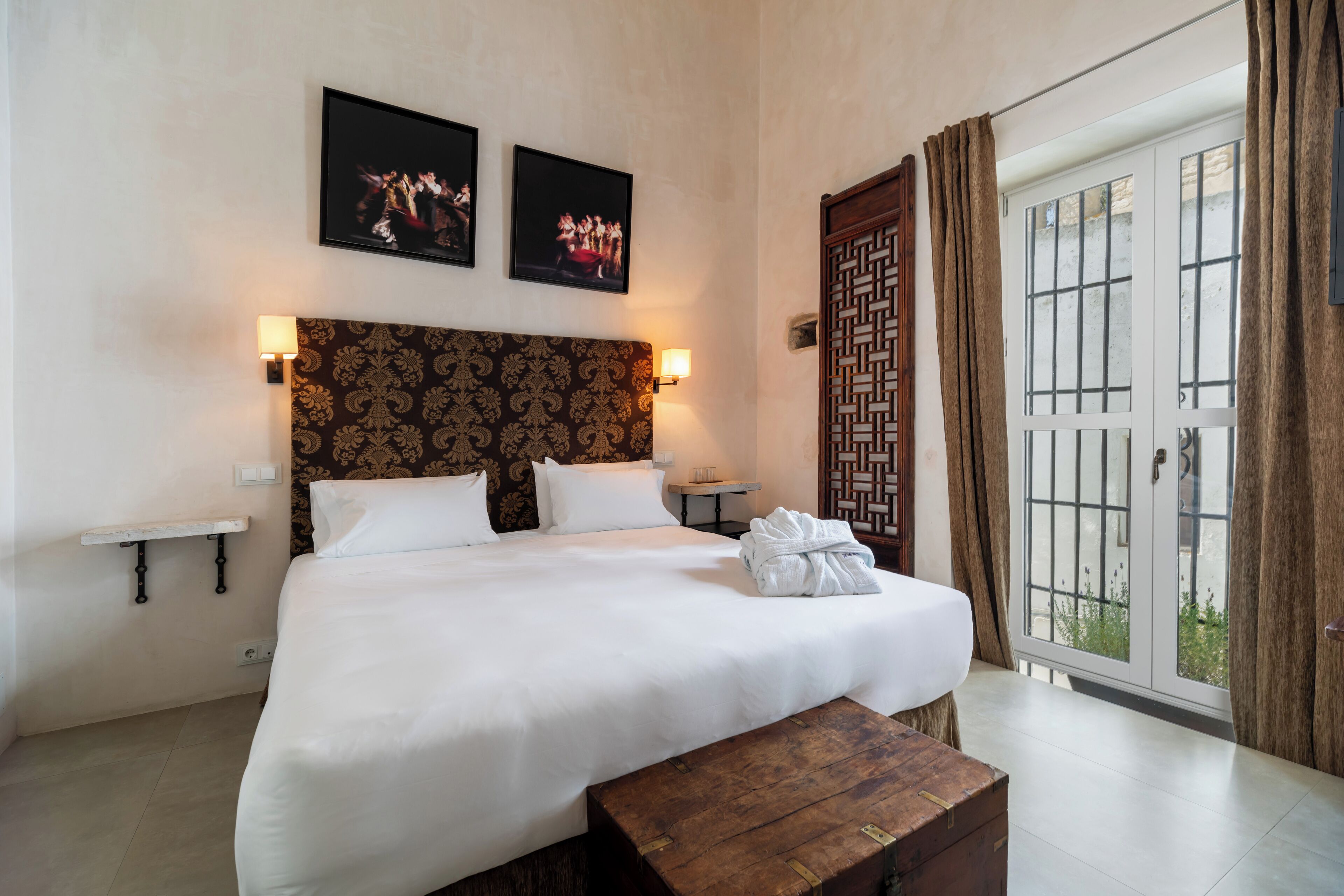 Een hotelkamer met een groot bed, een houten bankje, een nachtkastje, een wandlamp en een glazen schuifdeur naar een balkon.