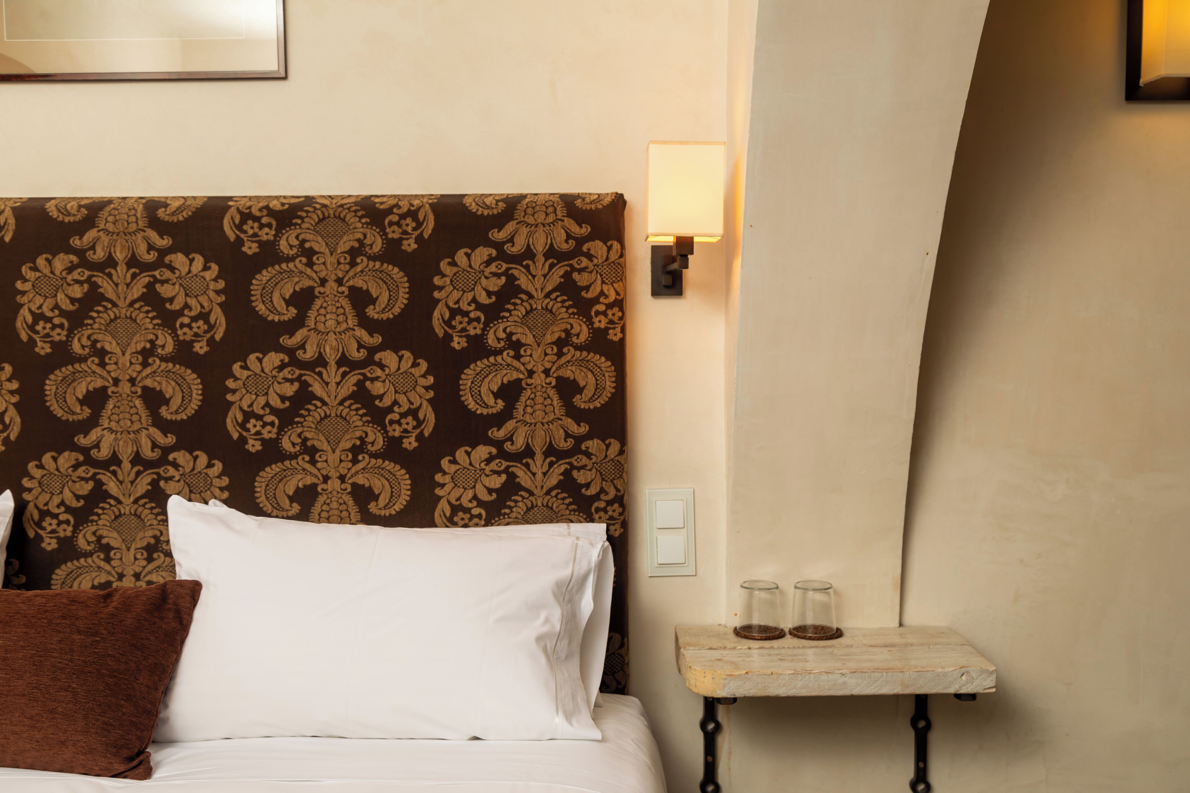 Quarto de hotel com cabeceira estampada, cama com roupa de cama branca, mesa de cabeceira pequena com dois copos e luminária fixada na parede.
